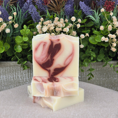 Three bars of Jarhead Aromatics Citrus + Patchouli Handmade Natural Tallow Artisan Soap, with the top bar featuring a marbled pink and cream swirl, are stacked in front of green foliage and purple flowers on a beige surface.