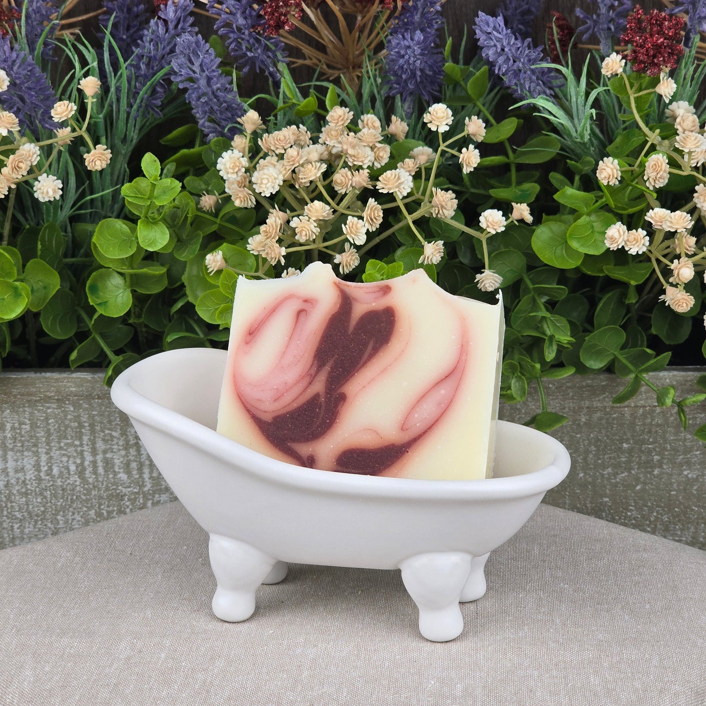 A bar of Jarhead Aromatics Citrus + Patchouli Handmade Natural Tallow Artisan Soap with a maroon swirl rests in a small white ceramic bathtub dish, surrounded by faux green leaves and purple and cream flowers in the background.