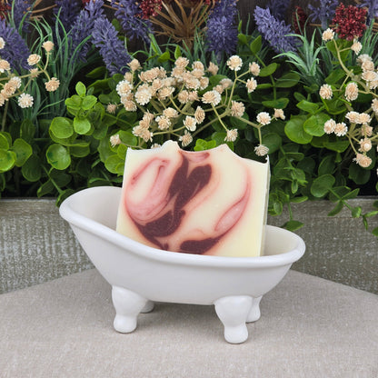 A bar of Jarhead Aromatics Citrus + Patchouli Handmade Natural Tallow Artisan Soap with a maroon swirl rests in a small white ceramic bathtub dish, surrounded by faux green leaves and purple and cream flowers in the background.
