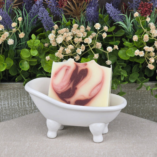 A bar of Jarhead Aromatics Citrus + Patchouli Handmade Natural Tallow Artisan Soap with a maroon swirl rests in a small white ceramic bathtub dish, surrounded by faux green leaves and purple and cream flowers in the background.