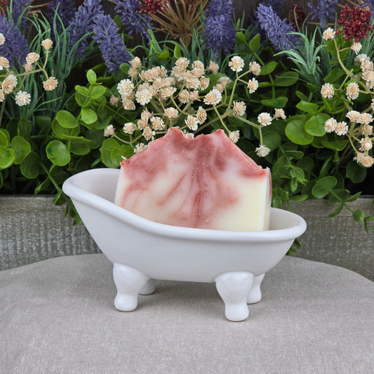 The Citrus + Tallow Handmade Artisan Soap by Jarhead Aromatics features pink and white swirls, displayed in a white clawfoot tub soap dish with greenery and purple flowers. Made with nourishing grass-fed beef tallow for a luxurious, all-natural cleanse.