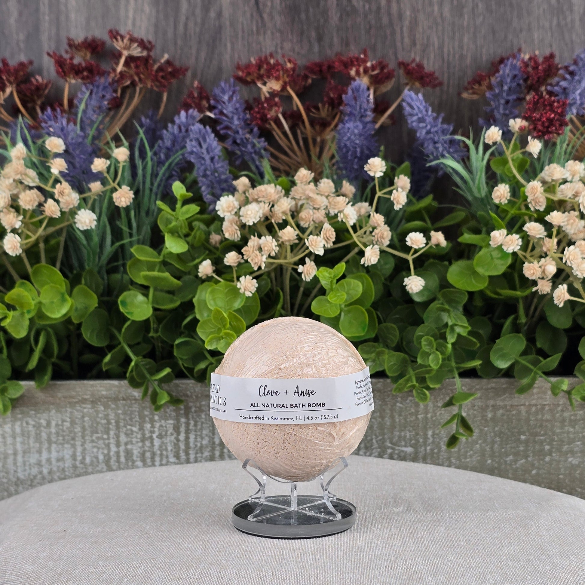 A Jarhead Aromatics Clove + Anise Bath Bomb - All Natural rests on a clear stand before lush green and purple faux plants, with a gray textured surface and wooden backdrop.