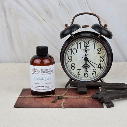 A brown bottle of Jarhead Aromatics’ Coastal Linen Diffuser Refill (Fresh Linen, Ozone & Sandalwood) sits beside a black alarm clock, metal keys, and a leather-bound book on a light surface with a marble background.