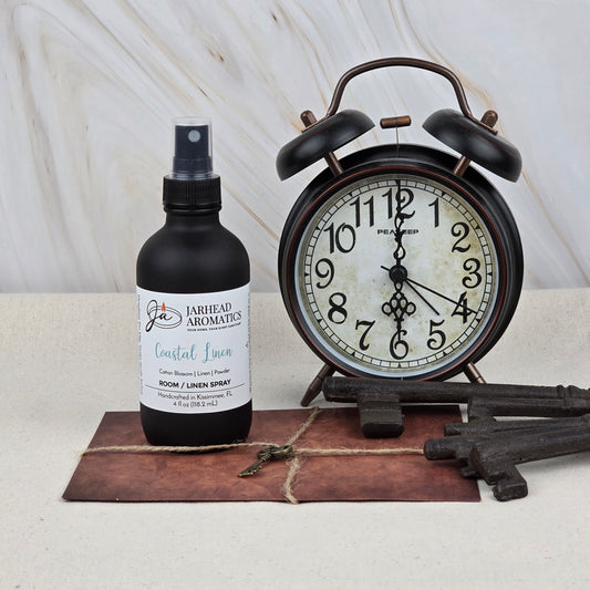 A vintage black alarm clock sits next to a bottle of Jarhead Aromatics Coastal Linen Room & Linen Spray, three large antique keys, and a brown envelope tied with twine on a light surface.