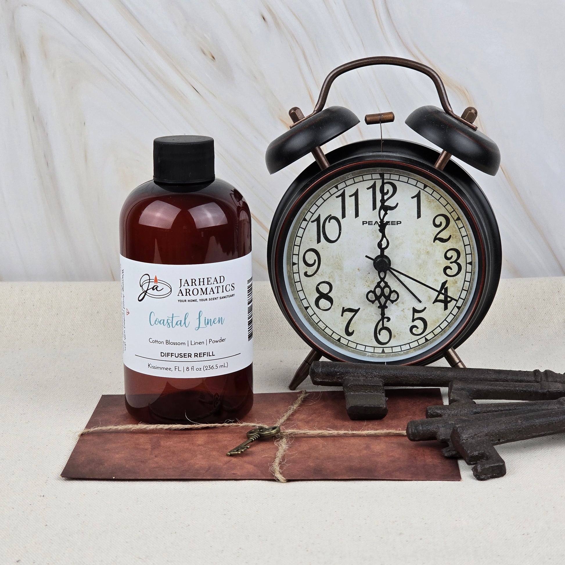 A bottle of Jarhead Aromatics Coastal Linen Diffuser Refill stands by a vintage black alarm clock at 12:44, with old keys on a brown envelope, filling the air with fresh linen, ozone, and sandalwood notes.