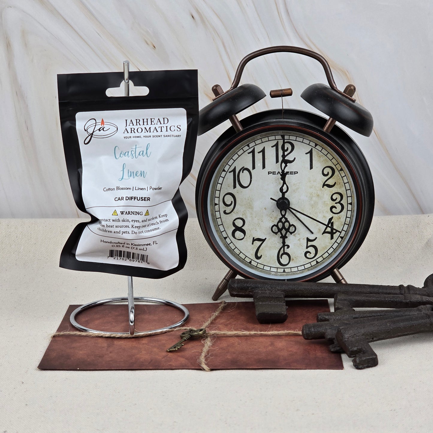 A package of Jarhead Aromatics Coastal Linen Car Diffuser | Fresh Linen & Ocean Breeze Car Air Freshener hangs on a metal stand beside a vintage black alarm clock, old-fashioned keys, and a brown leather notebook.
