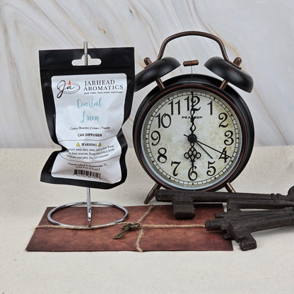 A package of Jarhead Aromatics Coastal Linen Car Diffuser | Fresh Linen & Ocean Breeze Car Air Freshener hangs on a metal stand beside a vintage black alarm clock, old-fashioned keys, and a brown leather notebook.