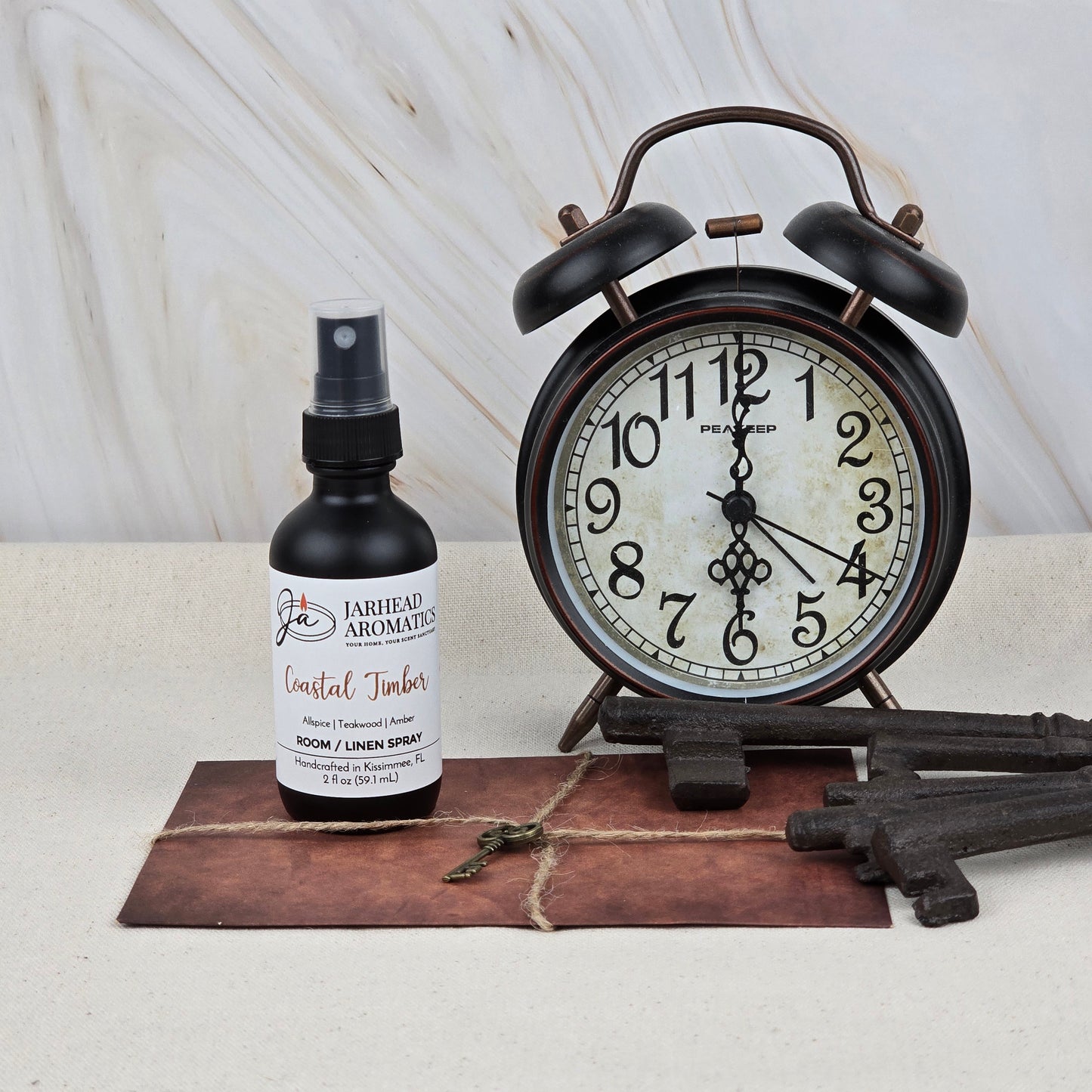 A black alarm clock showing 12:03 sits beside Jarhead Aromatics' Coastal Timber Room & Linen Spray (Teakwood, Leather & Amber). Vintage-style keys and a string-tied brown envelope rest in front, set against a marble wall for a chic home fragrance scene.