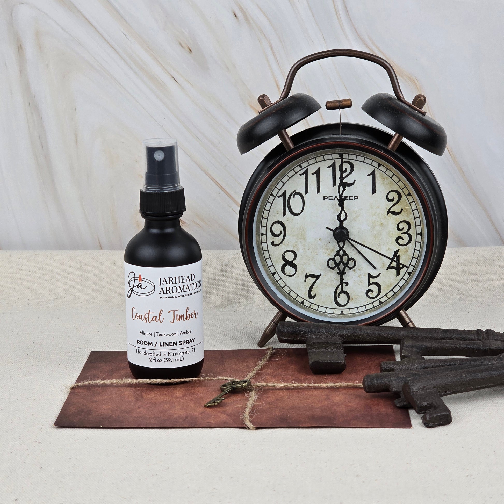 A black alarm clock showing 12:03 sits beside Jarhead Aromatics' Coastal Timber Room & Linen Spray (Teakwood, Leather & Amber). Vintage-style keys and a string-tied brown envelope rest in front, set against a marble wall for a chic home fragrance scene.