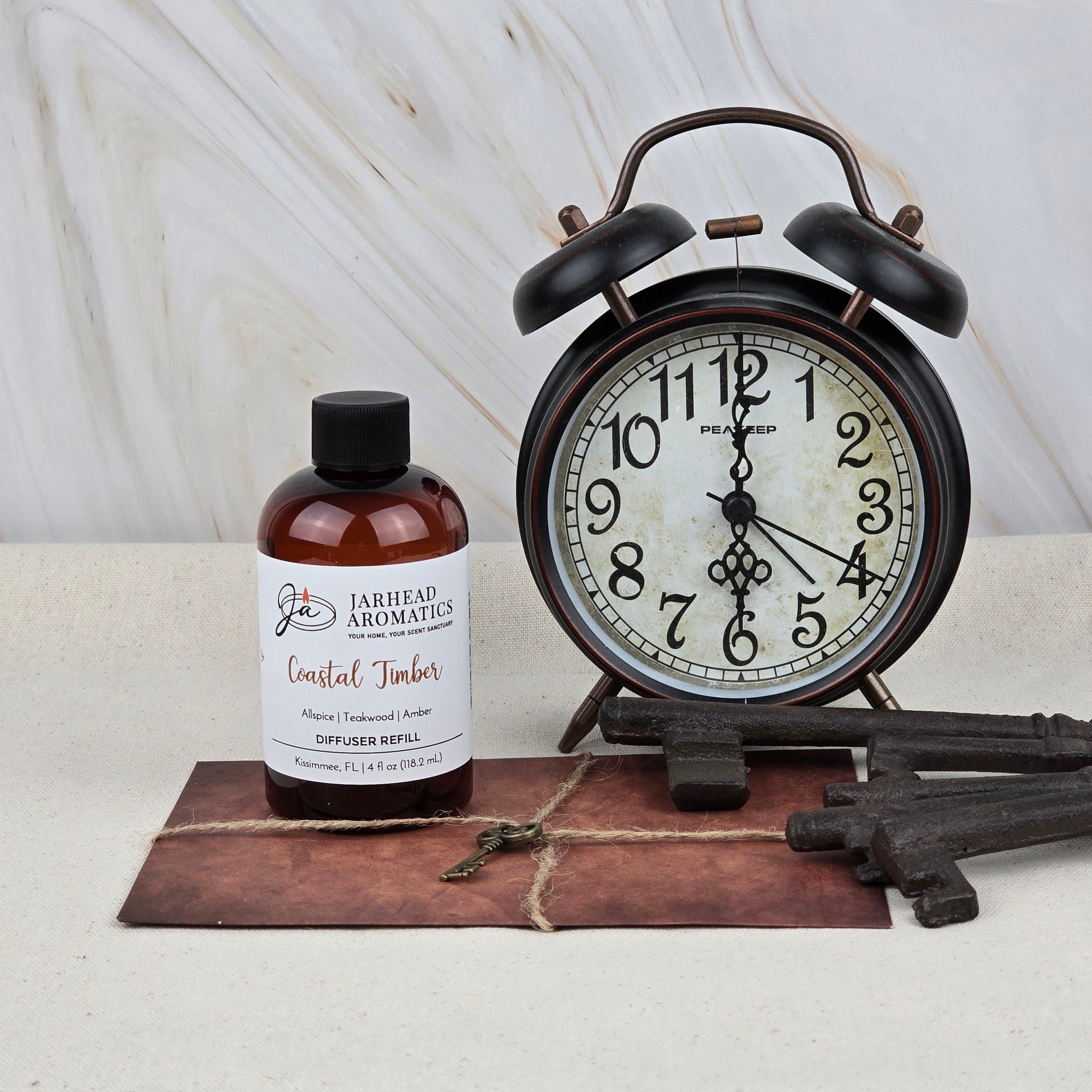 A bottle of Jarhead Aromatics Coastal Timber Diffuser Refill—featuring teakwood, leather, and amber notes—rests next to a vintage black alarm clock, old keys, and a brown leather-bound notebook on a light surface.