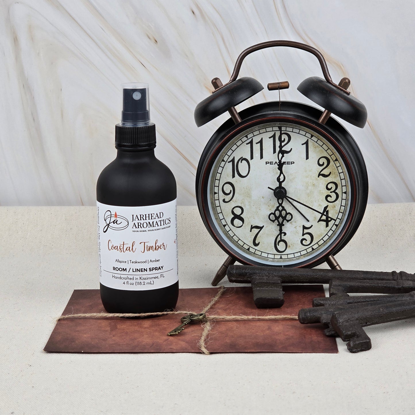 A bottle of Jarhead Aromatics Coastal Timber Room & Linen Spray | Teakwood, Leather & Amber rests beside a vintage black alarm clock and three antique keys on a brown envelope, all styled on a light surface for an elegant home fragrance touch.