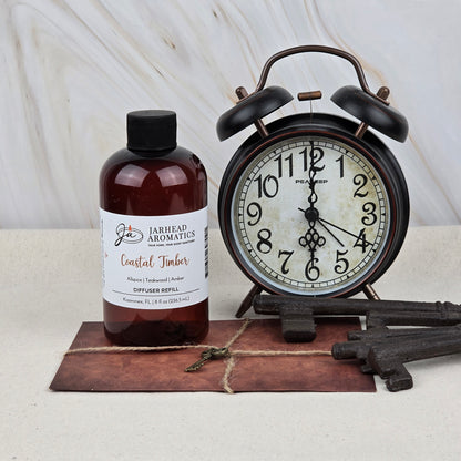 A brown bottle of Jarhead Aromatics Coastal Timber Diffuser Refill—Teakwood, Leather & Amber—sits by a black vintage alarm clock at 8:22, alongside three metal keys atop a twine-wrapped brown envelope.