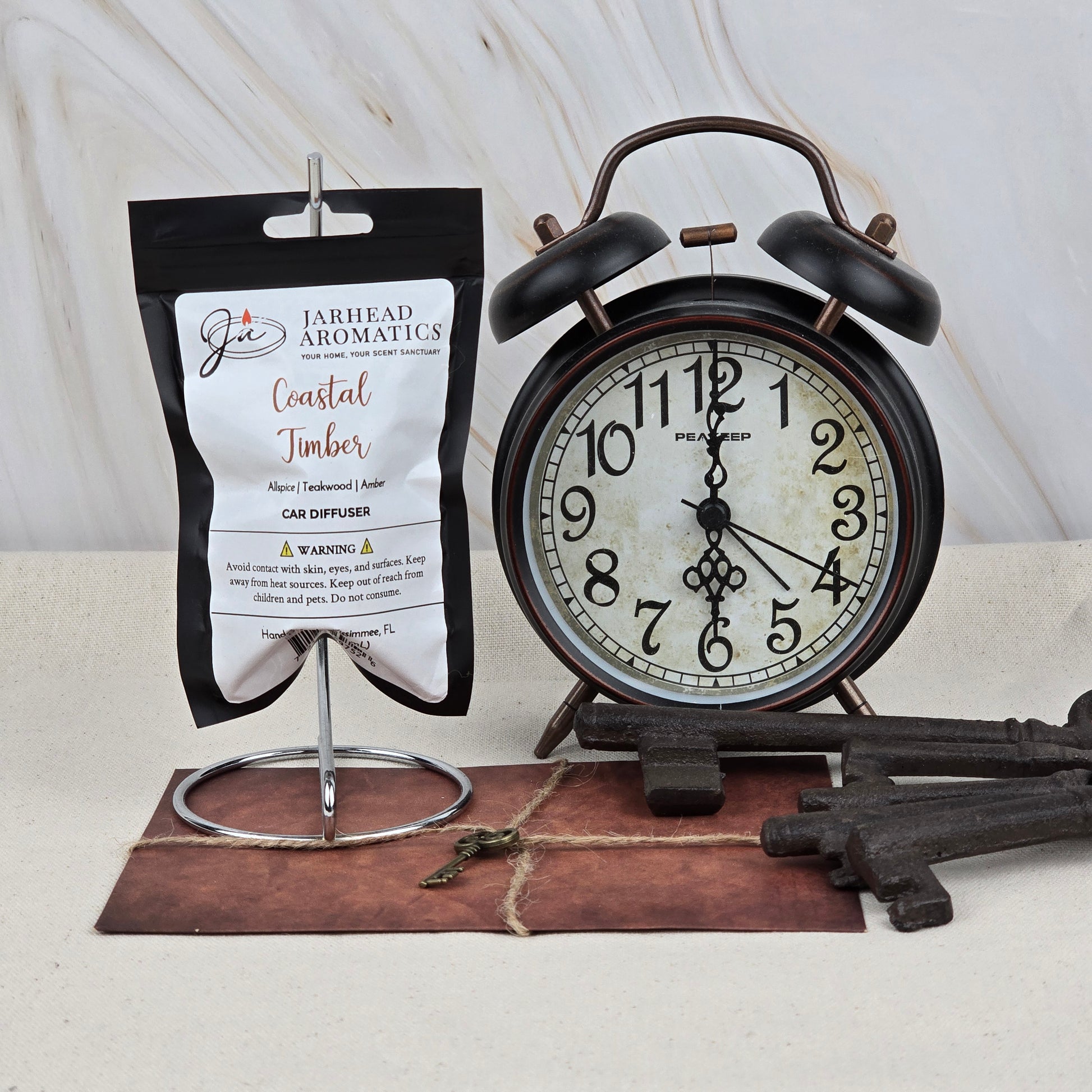 A black and white alarm clock sits beside a Coastal Timber Car Diffuser by Jarhead Aromatics. Large vintage keys and a small key rest on brown leather, highlighting this masculine teakwood, leather & amber car air freshener.
