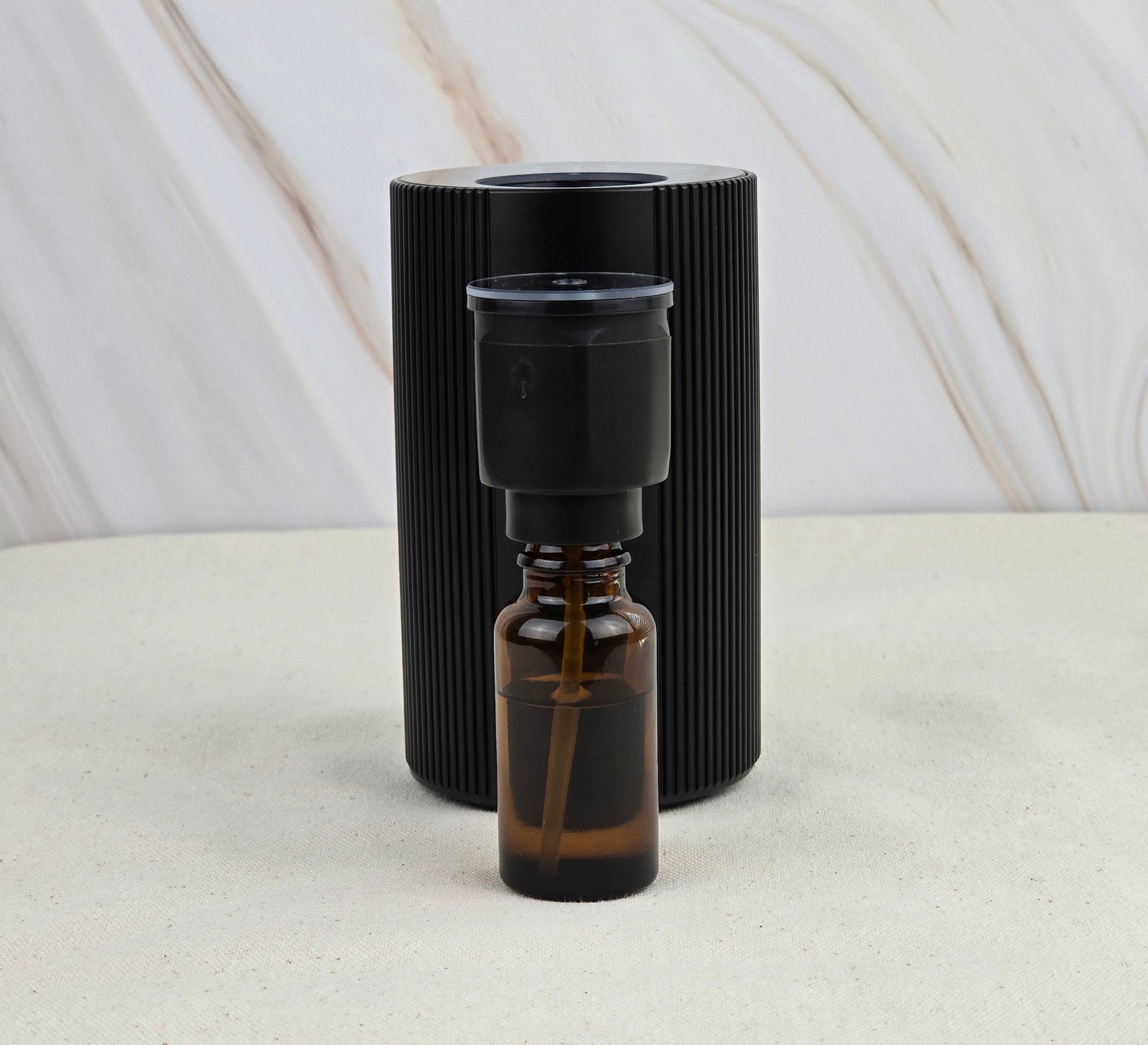 A small brown glass dropper bottle of earthy essential oils sits before the Jarhead Aromatics Cold Air Diffuser—Portable & Rechargeable, a black, vertically ridged cylinder—set against a neutral marbled background.