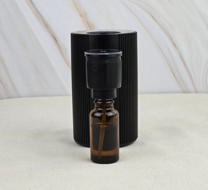 A small brown glass dropper bottle of earthy essential oils sits before the Jarhead Aromatics Cold Air Diffuser—Portable & Rechargeable, a black, vertically ridged cylinder—set against a neutral marbled background.