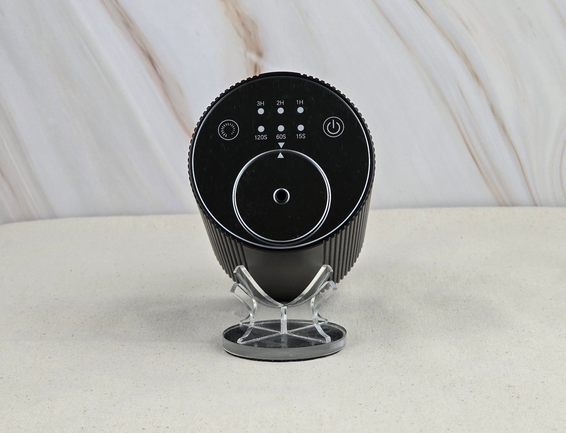 The Jarhead Aromatics Cold Air Diffuser - Portable & Rechargeable, a sleek black round device with buttons and a display, sits on a clear stand, bringing modern style to the light marble-patterned background.
