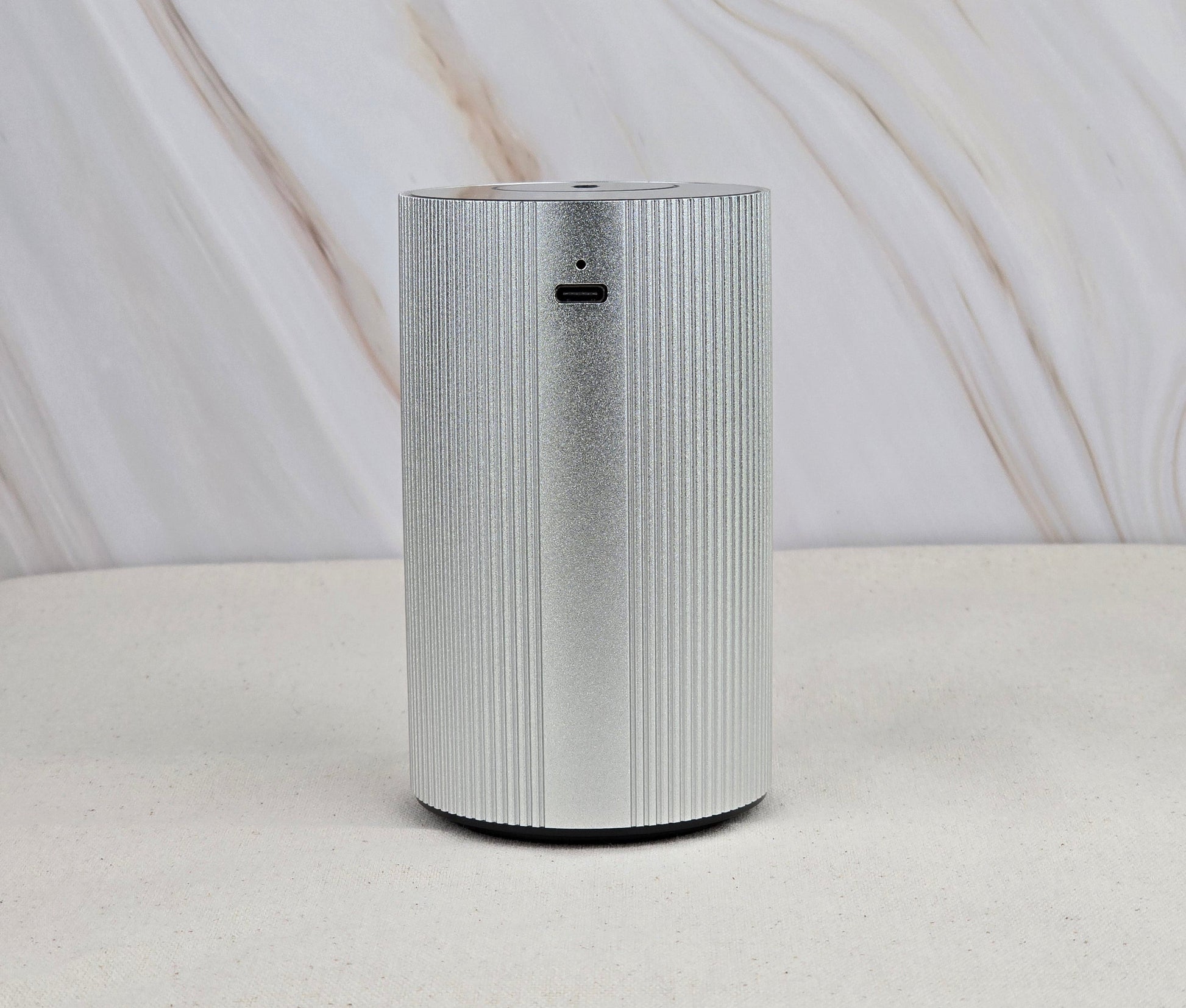 The Cold Air Diffuser - Portable & Rechargeable by Jarhead Aromatics, a sleek, cylindrical silver device with vertical grooves, sits on a light surface against a marble backdrop, bringing an earthy accent to the minimalist decor.