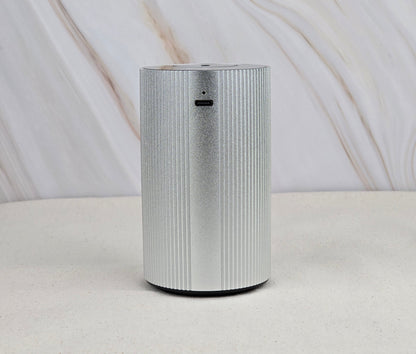 The Cold Air Diffuser - Portable & Rechargeable by Jarhead Aromatics, a sleek, cylindrical silver device with vertical grooves, sits on a light surface against a marble backdrop, bringing an earthy accent to the minimalist decor.