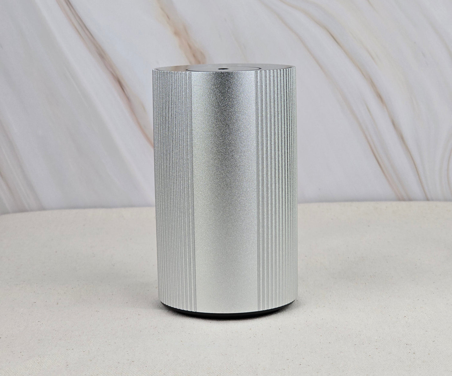 The Jarhead Aromatics Cold Air Diffuser - Portable & Rechargeable features a sleek silver cylindrical design with vertical ridges, displayed on a light surface and beige backdrop, evoking an earthy, woodsy ambiance inspired by essential oils.