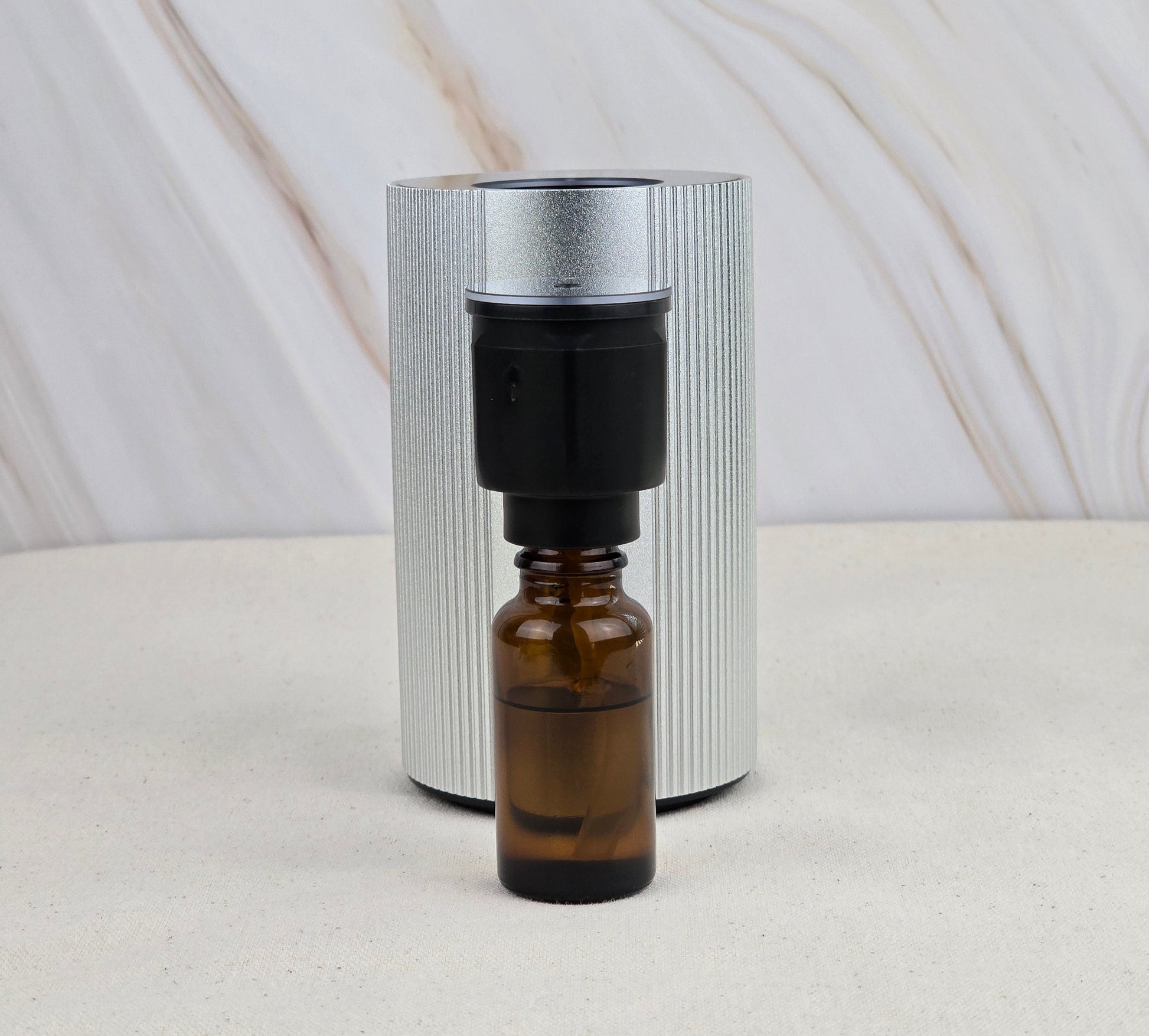 A compact Jarhead Aromatics Cold Air Diffuser - Portable & Rechargeable sits before a sleek silver cylinder on a marbled surface, ideal for dispersing your favorite earthy essential oils anywhere.