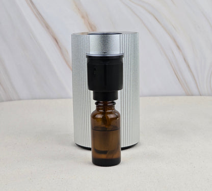 A compact Jarhead Aromatics Cold Air Diffuser - Portable & Rechargeable sits before a sleek silver cylinder on a marbled surface, ideal for dispersing your favorite earthy essential oils anywhere.
