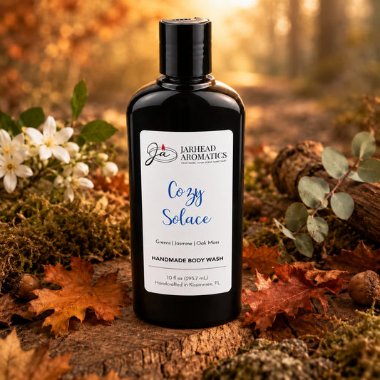 A bottle of Jarhead Aromatics Cozy Solace Handmade Body Wash, featuring notes of fresh greens, jasmine, and oak moss, rests on a forest floor scattered with autumn leaves and flowers beneath gentle sunlight.