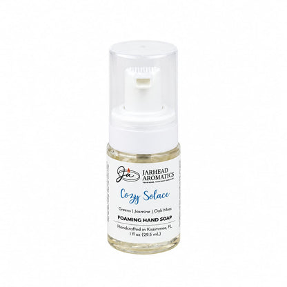 A small, clear plastic bottle of Cozy Solace Handmade Foaming Hand Soap with a white pump dispenser and a label featuring the botanical fragrance notes of greens, jasmine, and oak moss.