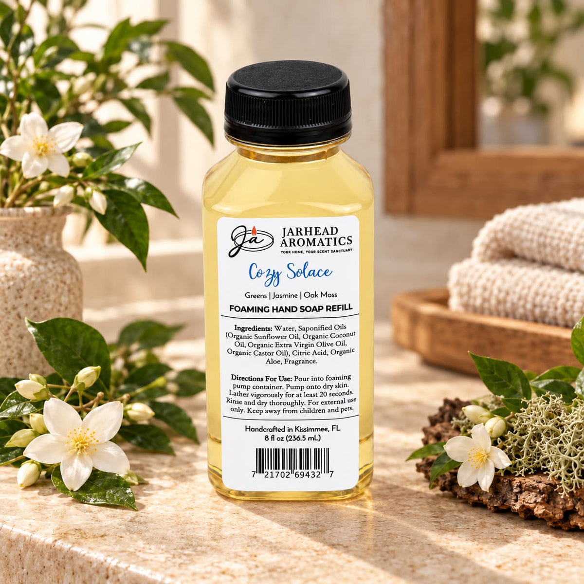 Clear bottle of Cozy Solace Handmade Foaming Hand Soap with a label detailing ingredients, directions, and scent notes, displayed on a marble countertop next to white flowers and moss.