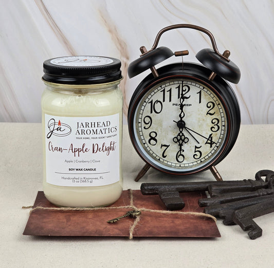 A Cran-Apple Delight Soy Wax Candle from Jarhead Aromatics sits by a vintage black alarm clock, with aged metal keys on a leather pouch atop a rustic white wood background.
