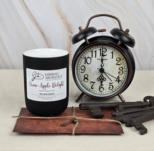 A black Cran-Apple Delight - Soy Wax Candle from Jarhead Aromatics, crafted with rich fragrance oil, sits beside a vintage black alarm clock on a beige surface. Several old-fashioned keys lie atop a rustic envelope tied with twine.