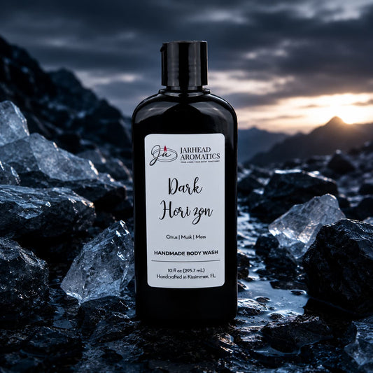 A bottle of Jarhead Aromatics Dark Horizon Handmade Body Wash, made with organic aloe and plant-based oils, stands among dark, glistening rocks with mountains and a moody sky in the background, evoking an air of masculine fragrance.