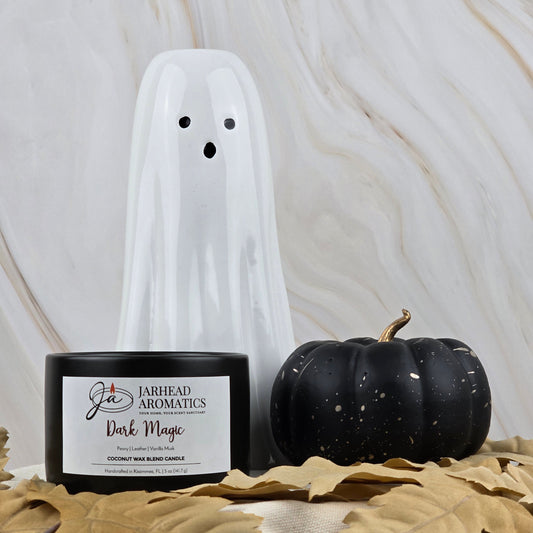 A white ceramic ghost, a black pumpkin, and Jarhead Aromatics’ Dark Magic Coconut Wax Blend Candle with vanilla musk notes sit on beige fabric and dried leaves before a marbled background.