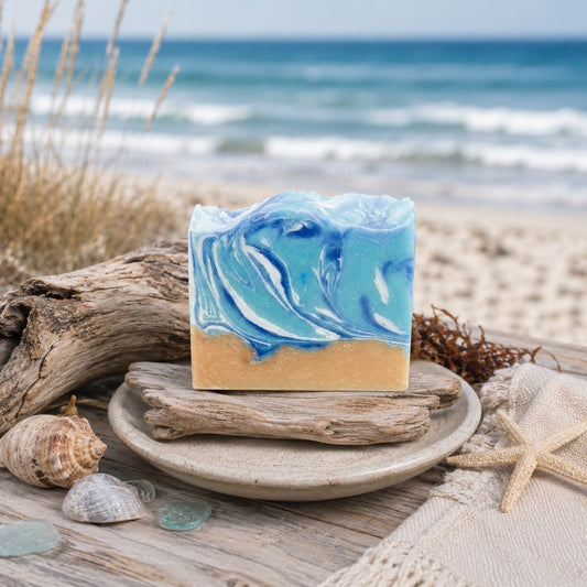 The Driftwood + Sea Salt Handmade Coconut Milk Artisan Soap by Jarhead Aromatics features blue and white swirls reminiscent of ocean waves, displayed on driftwood and surrounded by shells, sea glass, and sand with the beach and sea in the background.