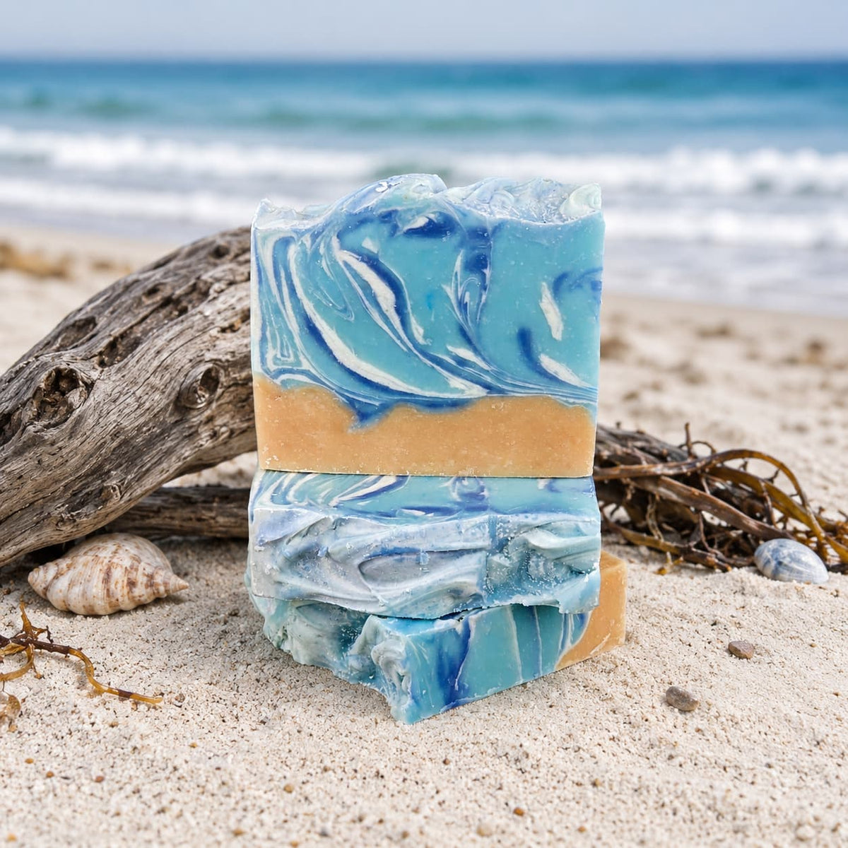 Two artisanal bars of Jarhead Aromatics Driftwood + Sea Salt Handmade Coconut Milk Artisan Soap, featuring blue, white, and tan swirls reminiscent of the coast, rest on a sandy beach near driftwood, seashells, and seaweed, with ocean waves and sky in the background.