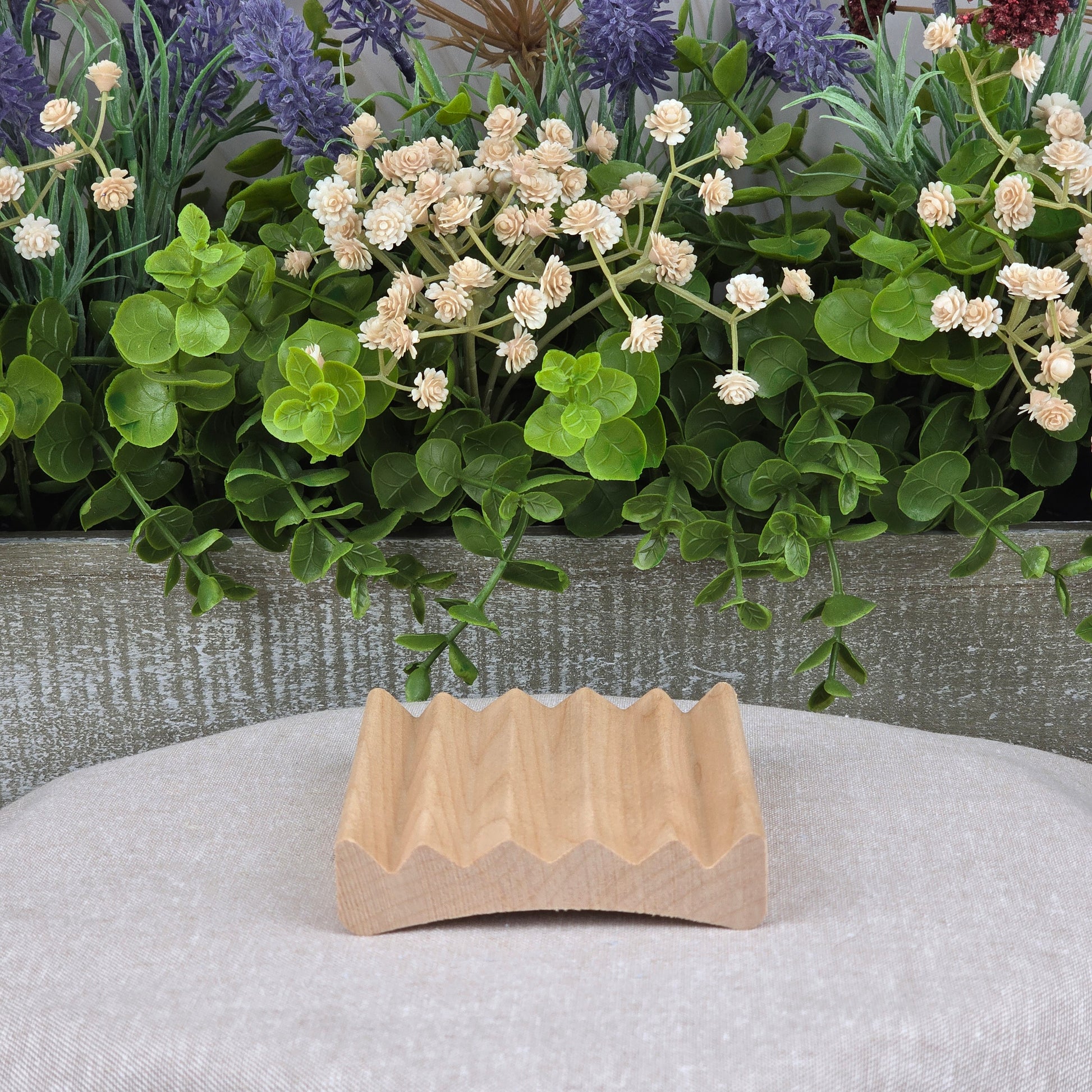The Elevation Style Wood Soap Dish by Jarhead Aromatics is a handcrafted, sustainable wooden soap saver with a wavy design, displayed on light gray fabric and accented by green leaves and white flowers in the background.