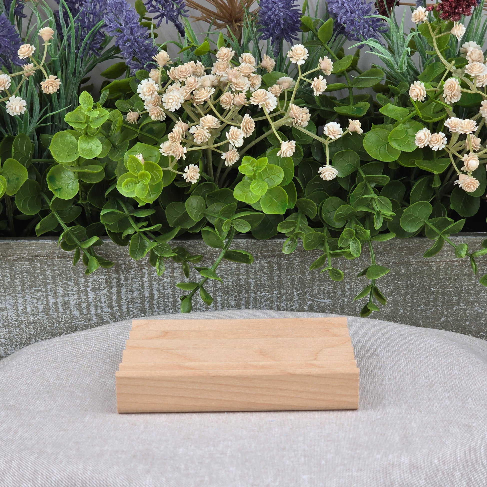 The Elevation Style Wood Soap Dish by Jarhead Aromatics, a handmade sustainable soap saver, rests on a light gray fabric surface with green leaves and white-purple artificial flowers in the background.