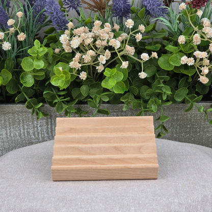 The Elevation Style Wood Soap Dish by Jarhead Aromatics, handcrafted from Red Alder with four grooves, rests on a gray cloth before faux green leaves and white-purple flowers—a sustainable, stylish soap saver for your bathroom.
