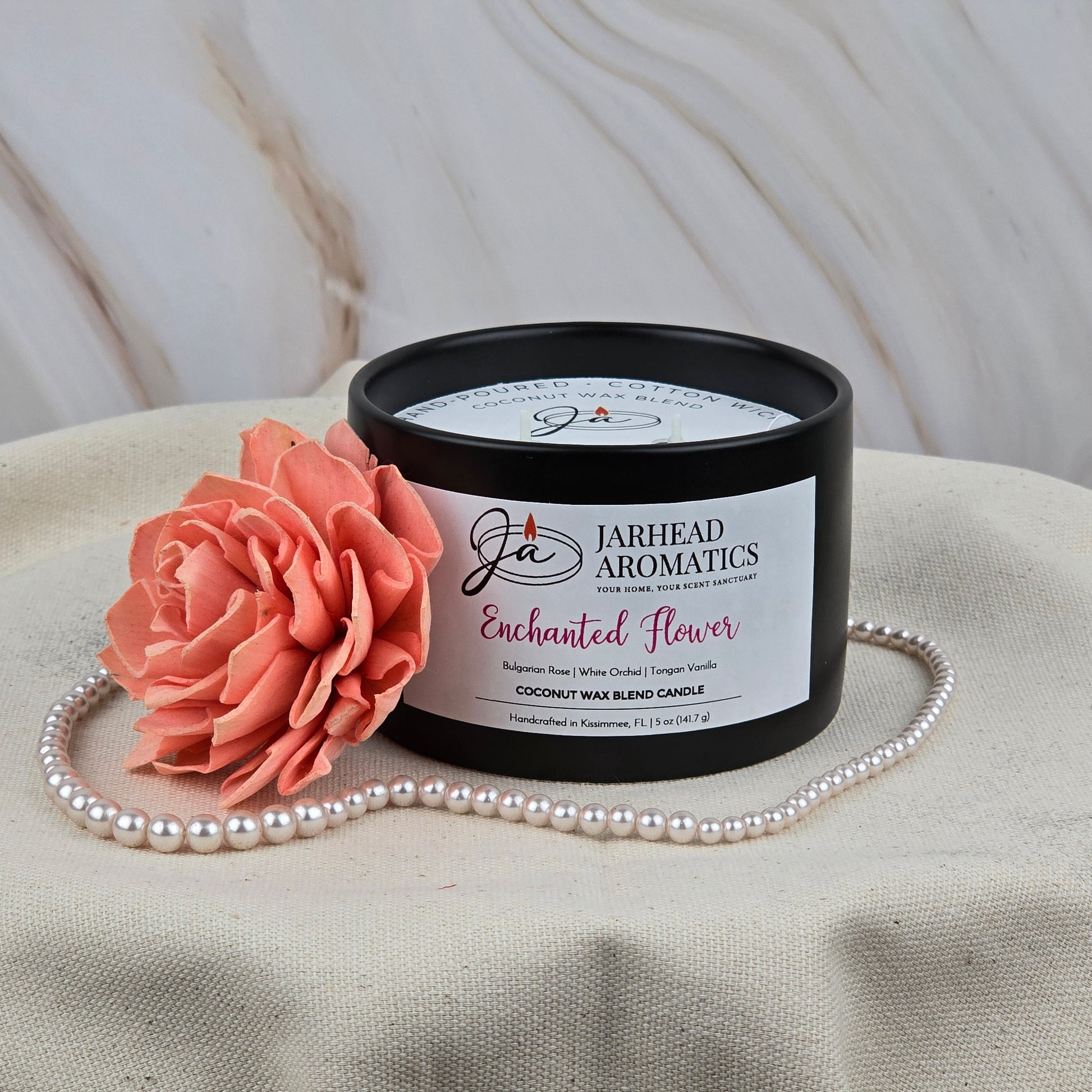 A Jarhead Aromatics Enchanted Flower Coconut Wax Blend Candle—featuring rose, orchid & vanilla—rests on cream fabric beside a pink artificial flower and pearl necklace, set against a marble background for a touch of luxury fragrance.