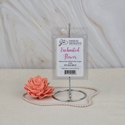 The Enchanted Flower Soy Wax Melt by Jarhead Aromatics, scented with rose orchid and vanilla, is displayed upright on a metal stand. In front, a pink flower candle and pearls rest on beige, evoking its floral aroma.