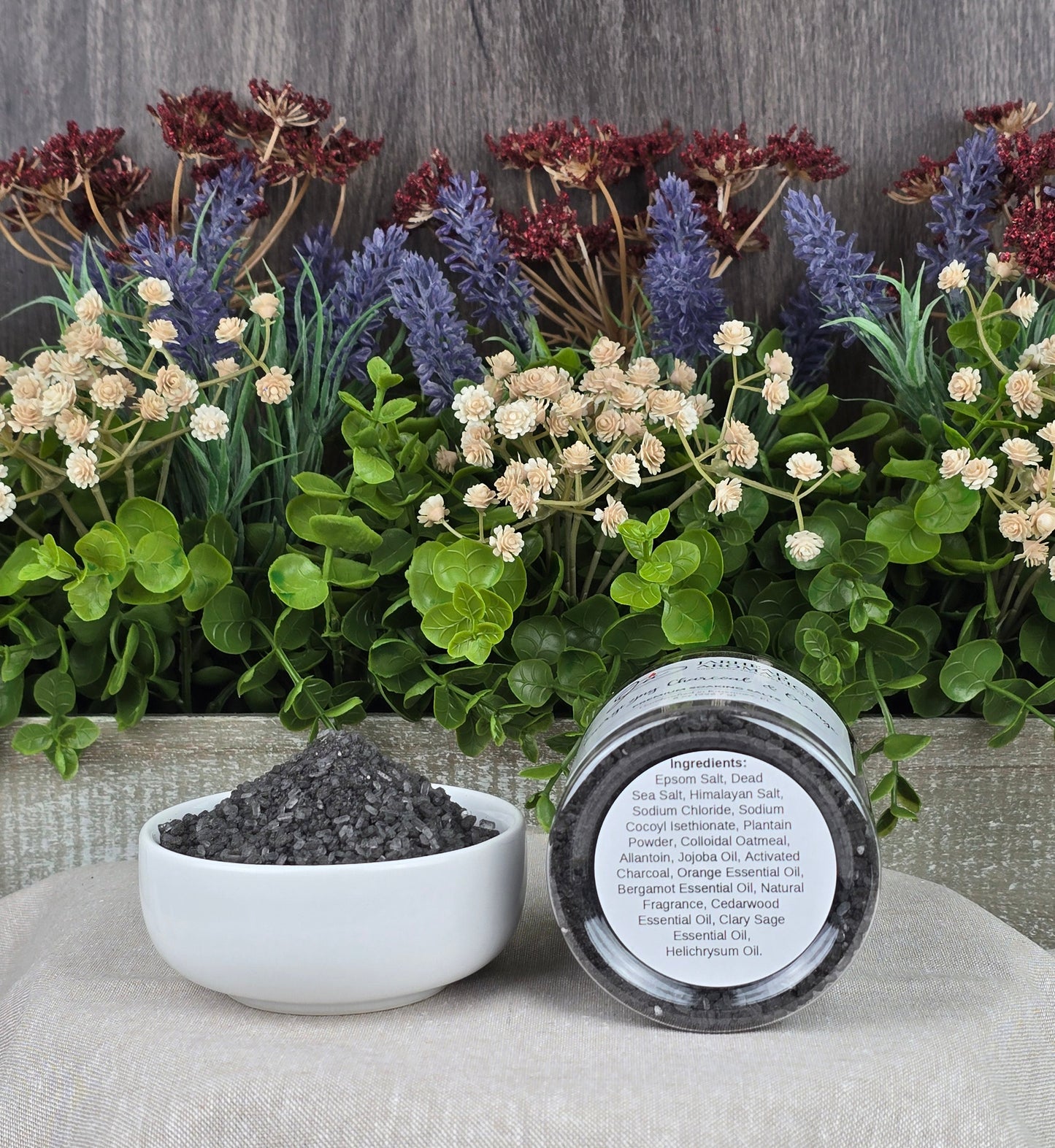 A small bowl of dark granules sits on a beige cloth beside a jar labeled Energizing Charcoal & Orange Premium Soaking Bath Salts by Jarhead Aromatics. Decorative green leaves and purple and red flowers form a serene backdrop, perfect for detox and skin nourishment.