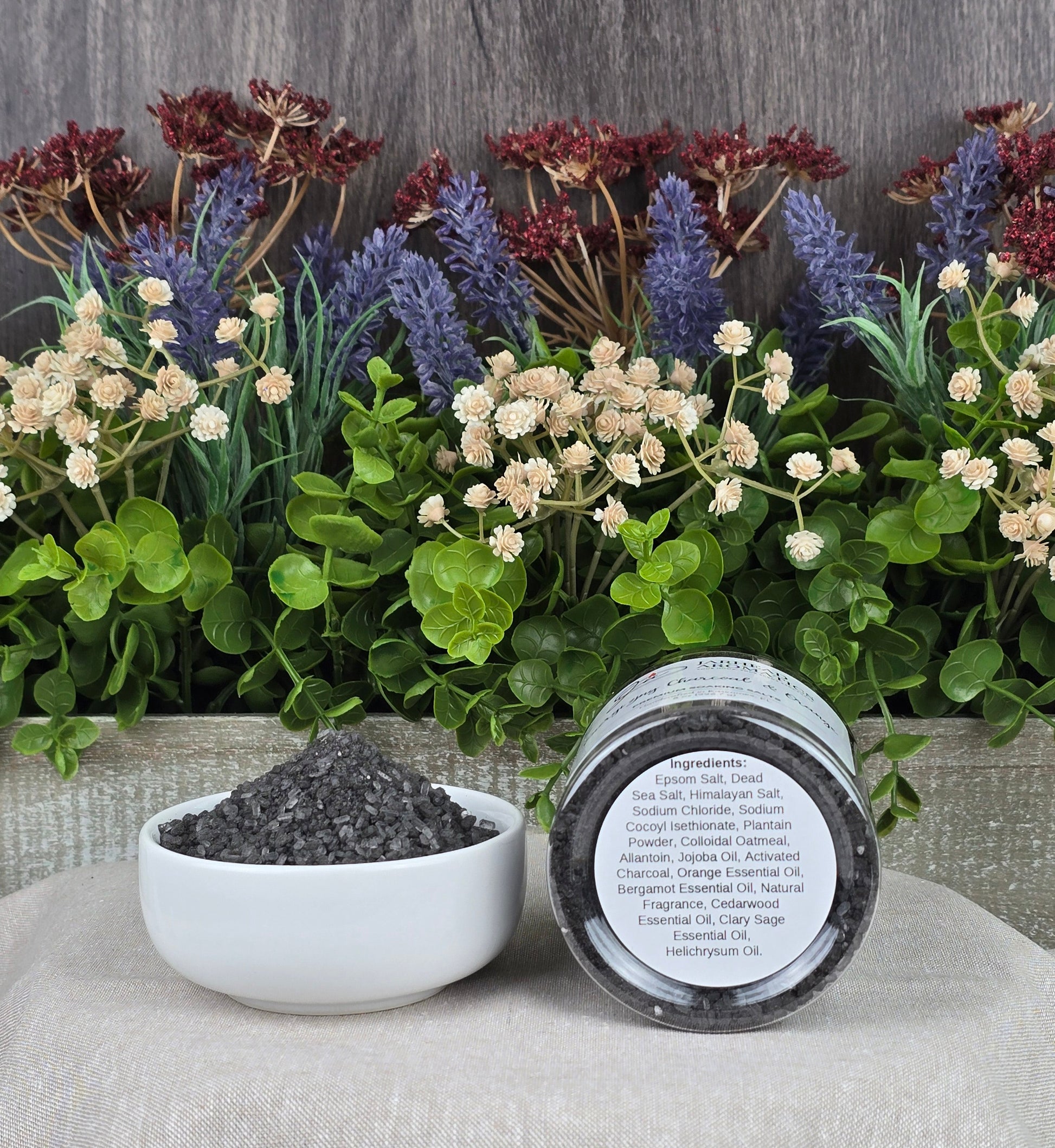 A small bowl of dark granules sits on a beige cloth beside a jar labeled Energizing Charcoal & Orange Premium Soaking Bath Salts by Jarhead Aromatics. Decorative green leaves and purple and red flowers form a serene backdrop, perfect for detox and skin nourishment.