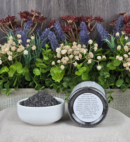 A small bowl of dark granules sits on a beige cloth beside a jar labeled Energizing Charcoal & Orange Premium Soaking Bath Salts by Jarhead Aromatics. Decorative green leaves and purple and red flowers form a serene backdrop, perfect for detox and skin nourishment.