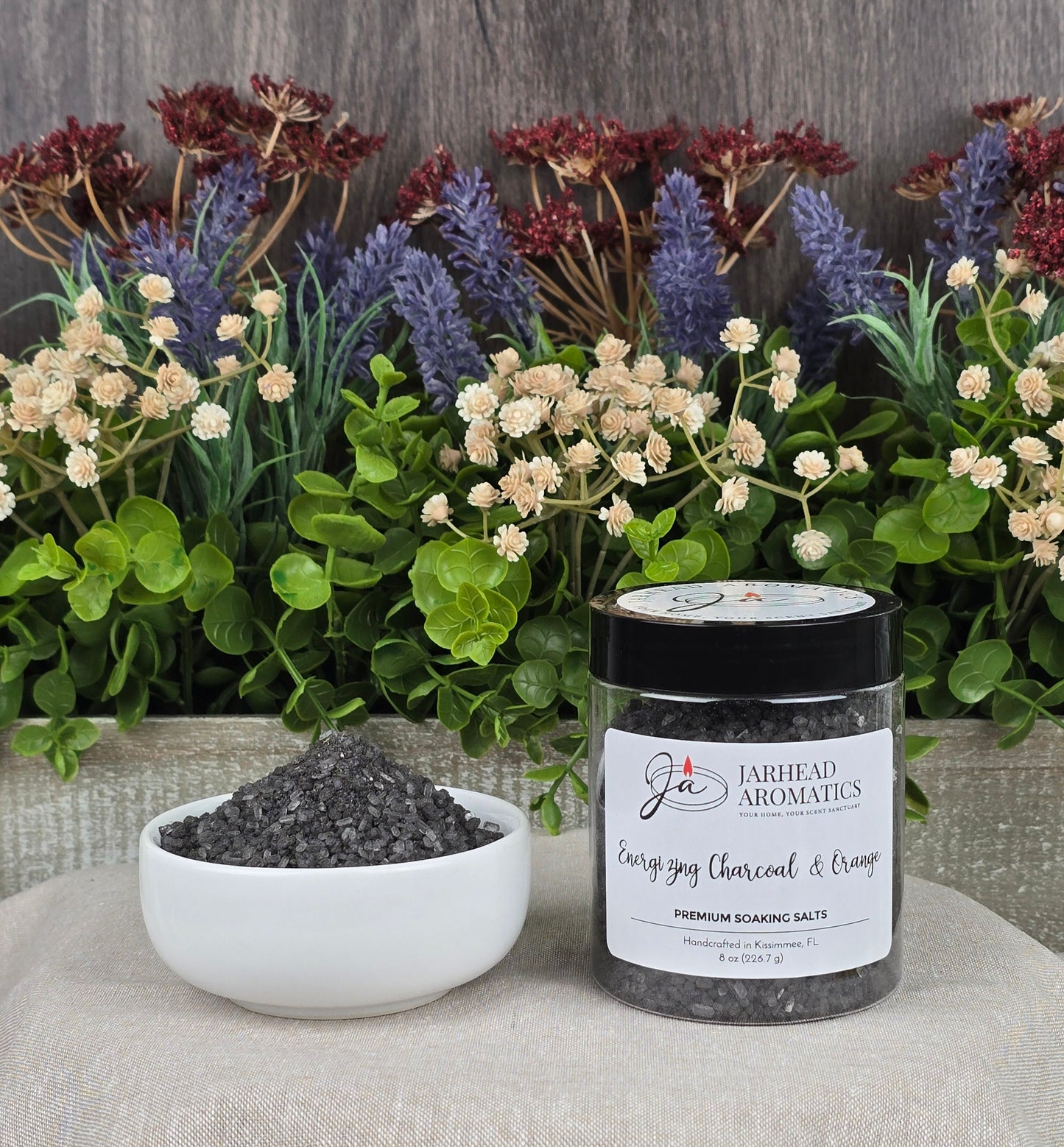 A jar of Jarhead Aromatics Energizing Charcoal & Orange Premium Soaking Bath Salts, known for skin nourishment, sits beside a white bowl filled with black salts amidst flowers and greenery on patterned fabric.