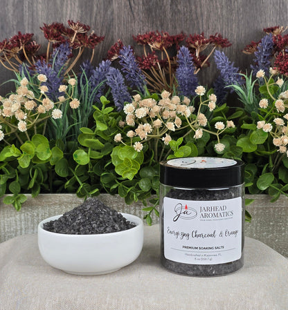 A jar of Jarhead Aromatics Energizing Charcoal & Orange Premium Soaking Bath Salts, known for skin nourishment, sits beside a white bowl filled with black salts amidst flowers and greenery on patterned fabric.