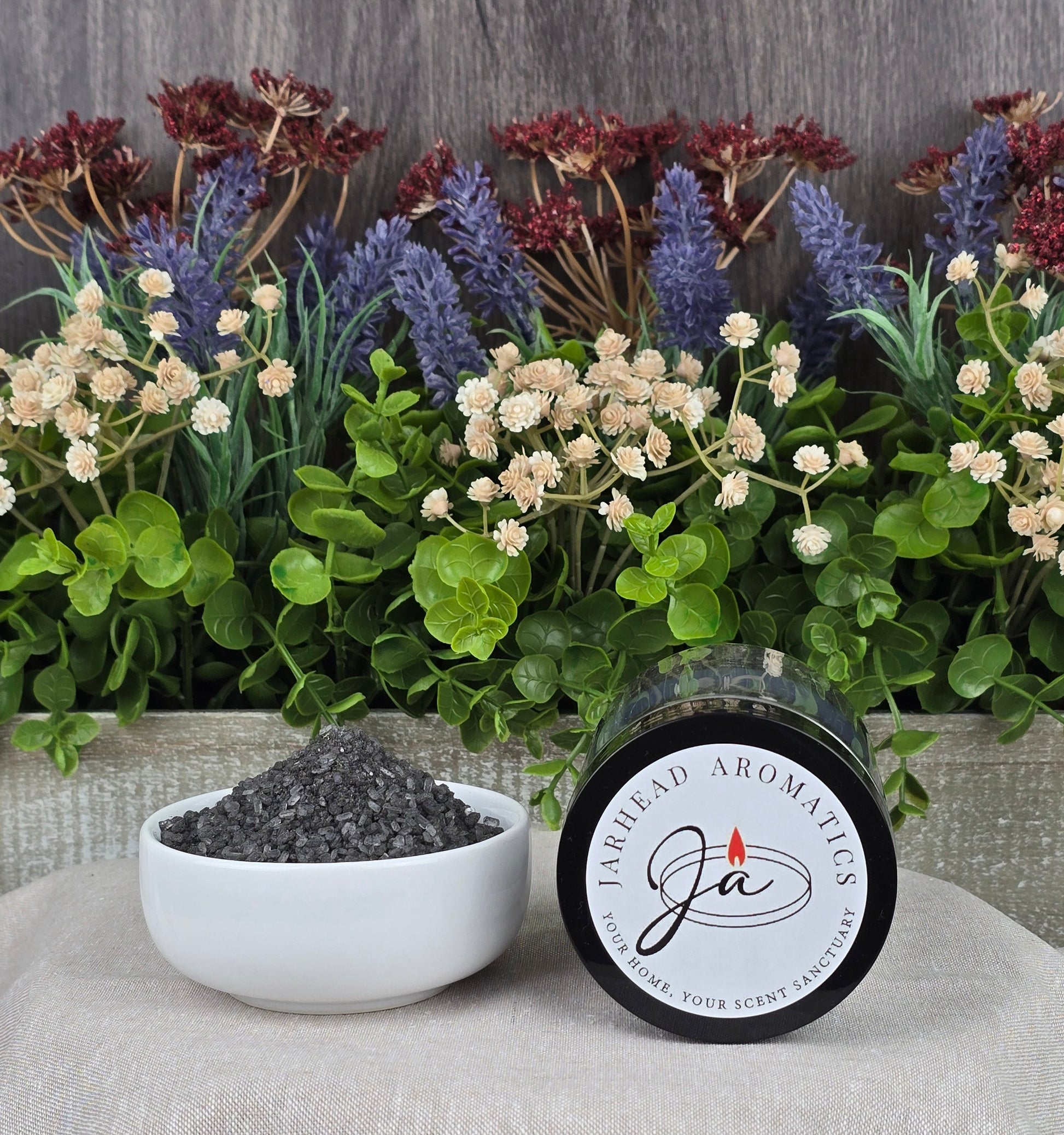 A jar of Energizing Charcoal & Orange Premium Soaking Bath Salts - All Natural by Jarhead Aromatics spills into a white bowl surrounded by lush green leaves and dried lavender, promising detoxification and skin nourishment.
