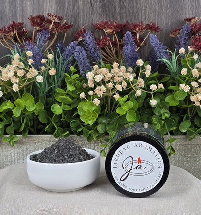 A jar of Energizing Charcoal & Orange Premium Soaking Bath Salts - All Natural by Jarhead Aromatics spills into a white bowl surrounded by lush green leaves and dried lavender, promising detoxification and skin nourishment.