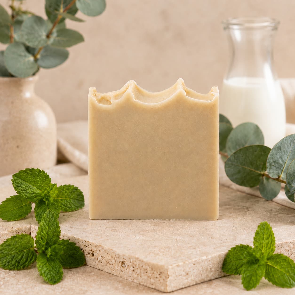 A bar of Jarhead Aromatics’ Eucalyptus + Mint Handmade Natural Goat Milk Artisan Soap, featuring a wavy top and a soothing beige hue, rests on a stone surface surrounded by fresh green mint leaves, a leafy vase of eucalyptus, and a glass bottle of milk in the background.