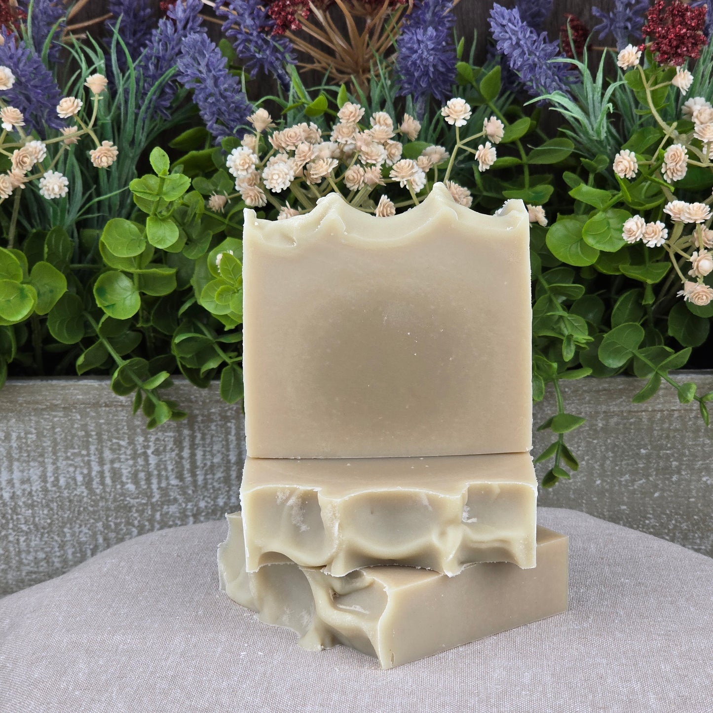 Three Jarhead Aromatics Eucalyptus + Mint Handmade Natural Goat Milk Artisan Soaps are stacked with the top bar upright, surrounded by green leaves and dried flowers for a refreshing touch.
