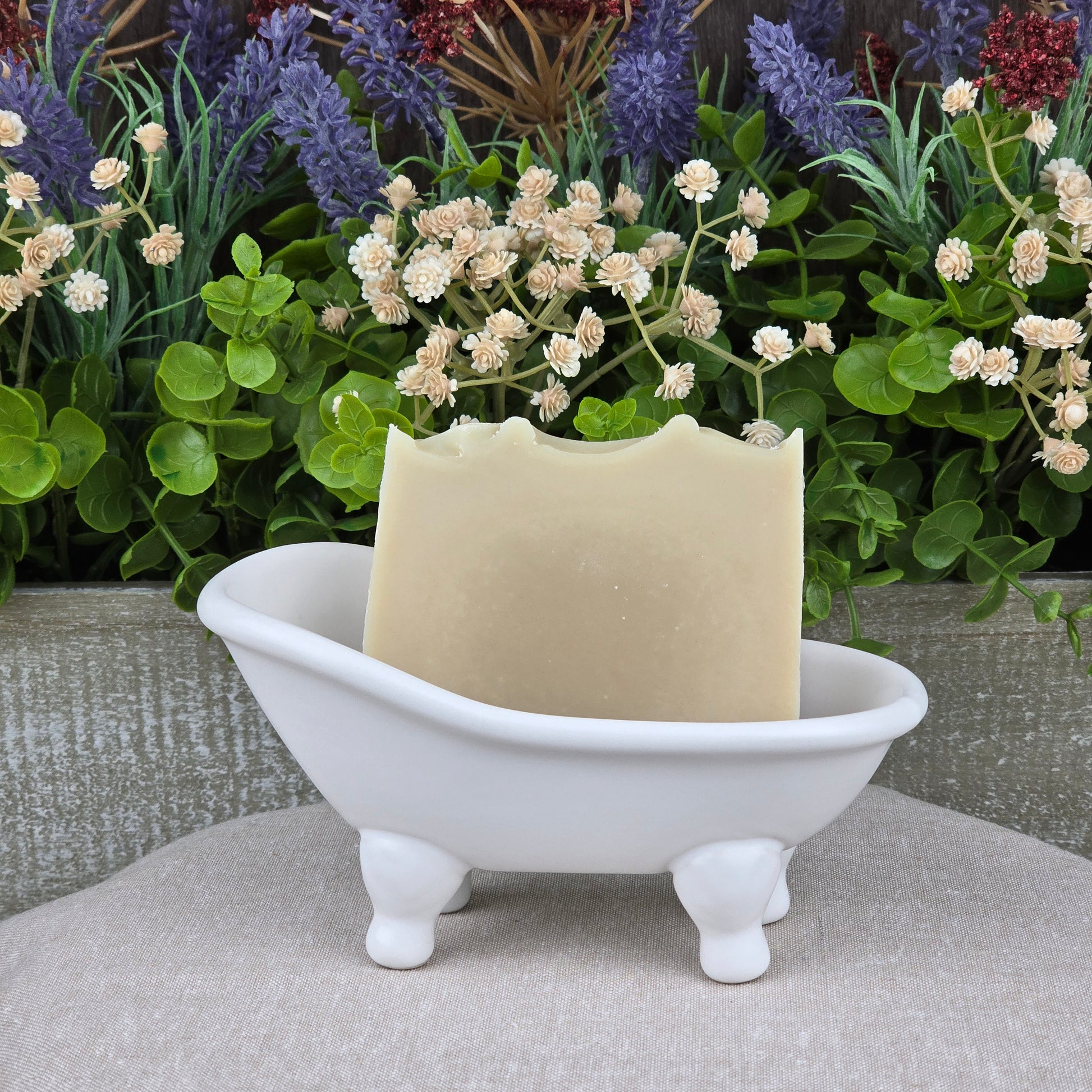 A bar of Jarhead Aromatics Eucalyptus + Mint Handmade Natural Goat Milk Artisan Soap rests in a small white ceramic bathtub dish, with green leaves and purple and white flowers in the background.