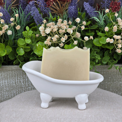A bar of Jarhead Aromatics Eucalyptus + Mint Handmade Natural Goat Milk Artisan Soap rests in a small white ceramic bathtub dish, with green leaves and purple and white flowers in the background.